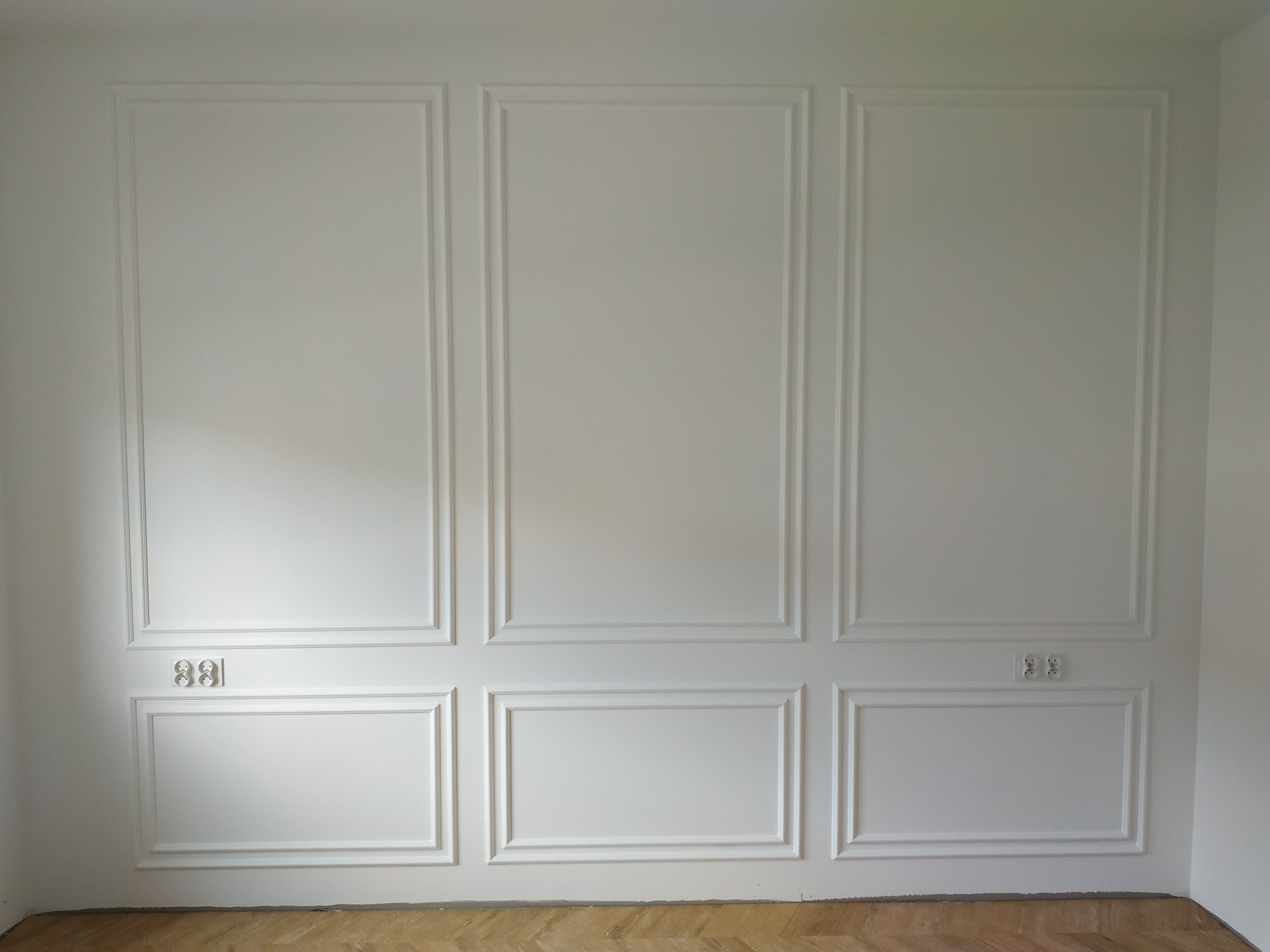 Biała ściana z dekoracyjnymi panelami ściennymi i gniazdkami elektrycznymi, parkiet na podłodze.