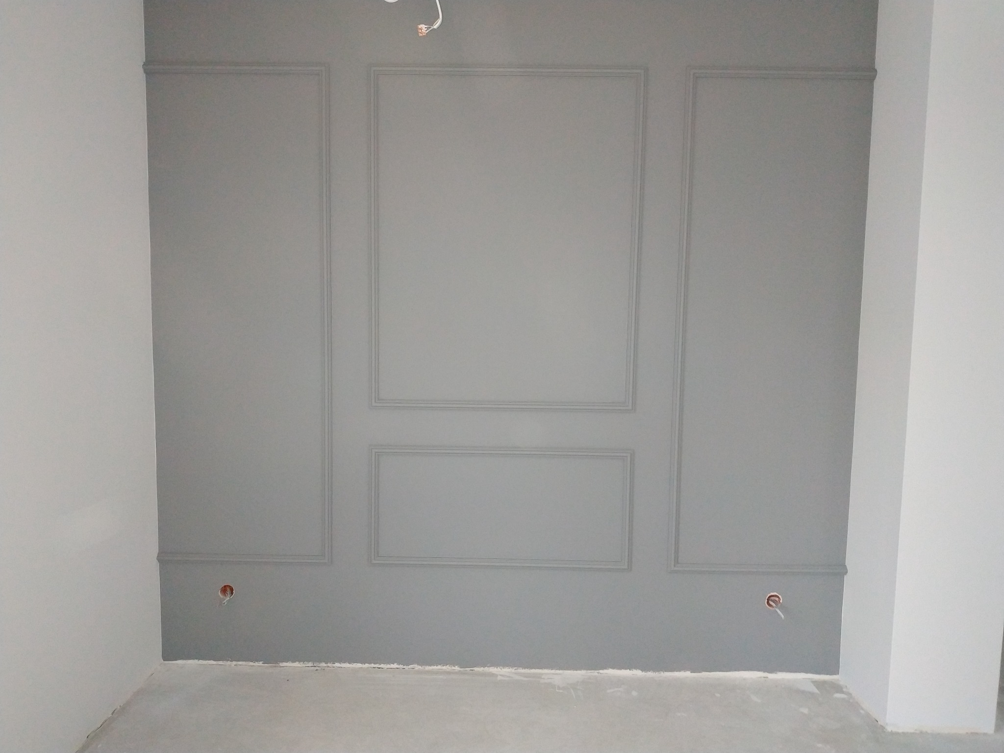 Szara ściana z dekoracyjnymi panelami w kształcie prostokątów i kwadratów, widoczne gniazdka elektryczne, surowy beton na podłodze, stan surowy.