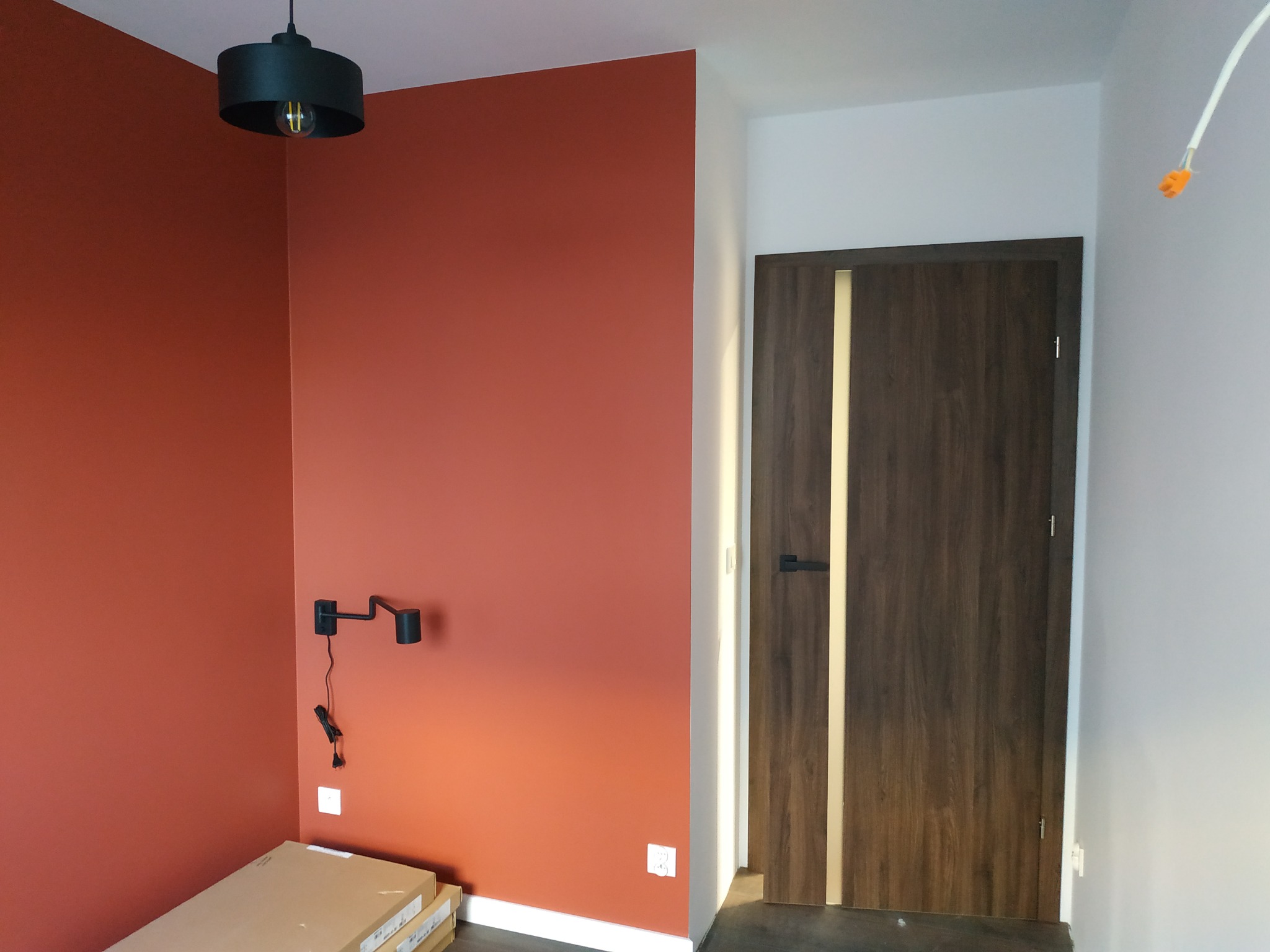 Ściana pomalowana na rdzawy kolor z zamontowanym czarnym kinkietem, widoczna drewniana drzwi z jasnym paskiem, lampa sufitowa i kartony na podłodze.