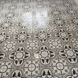 Ułożona podłoga z płytek ceramicznych w geometryczny wzór w odcieniach szarości i bieli, widoczne fugi i odbicie światła na powierzchni.