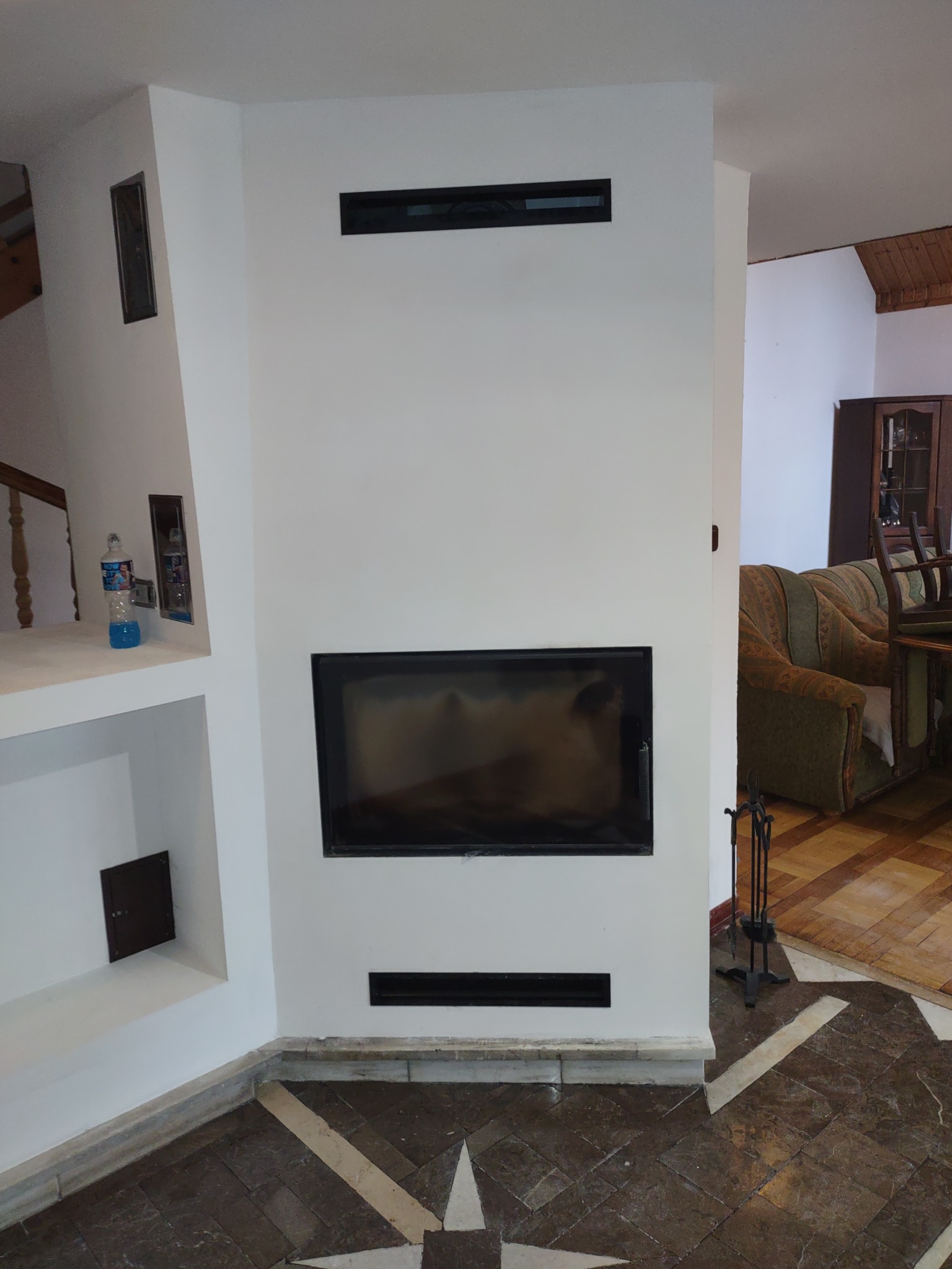 Biała ściana z wnękami i czarnym wkładem kominkowym, widoczne detale wykończenia podłogi i elementy wystroju wnętrza w tle.