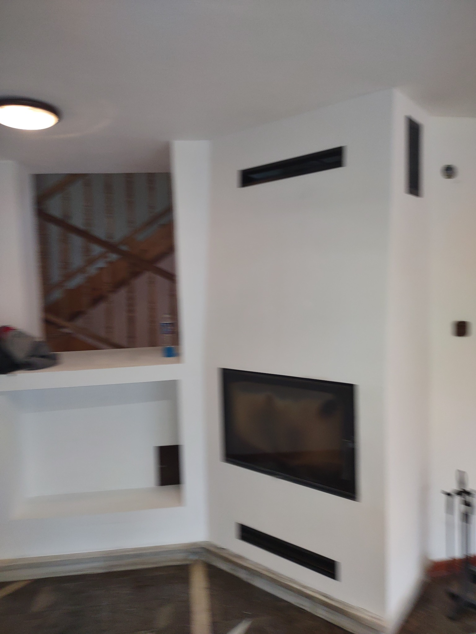 Biała ściana z wnęką i kominkiem z czarnymi kratkami wentylacyjnymi oraz półkami, widoczne elementy wykończenia podłogi.