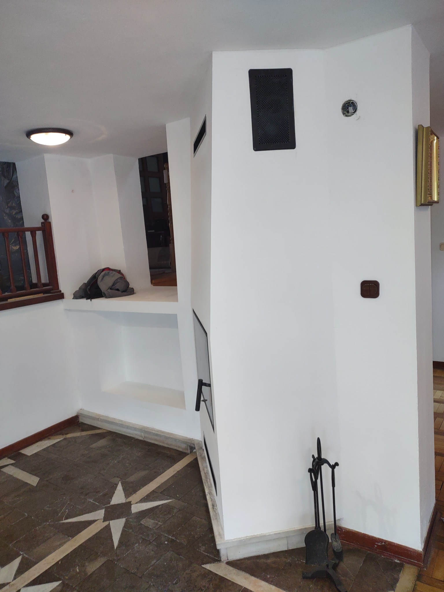 Białe wnętrze z dekoracyjną podłogą w gwiazdę, z wnęką i kominkiem z akcesoriami, widoczne elementy wykończenia i instalacje.