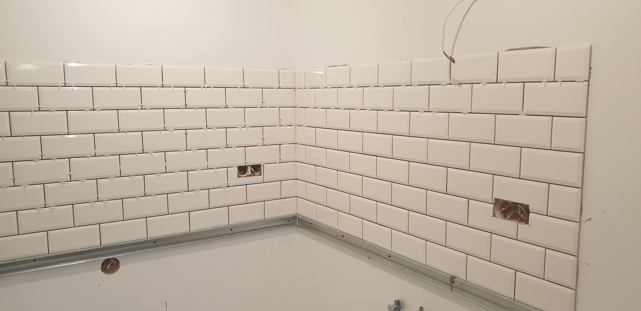 Układanie białych, prostokątnych płytek na ścianie z widocznymi krzyżykami dystansowymi, metalową listwą przypodłogową i otworami na gniazdka elektryczne w trakcie remontu.