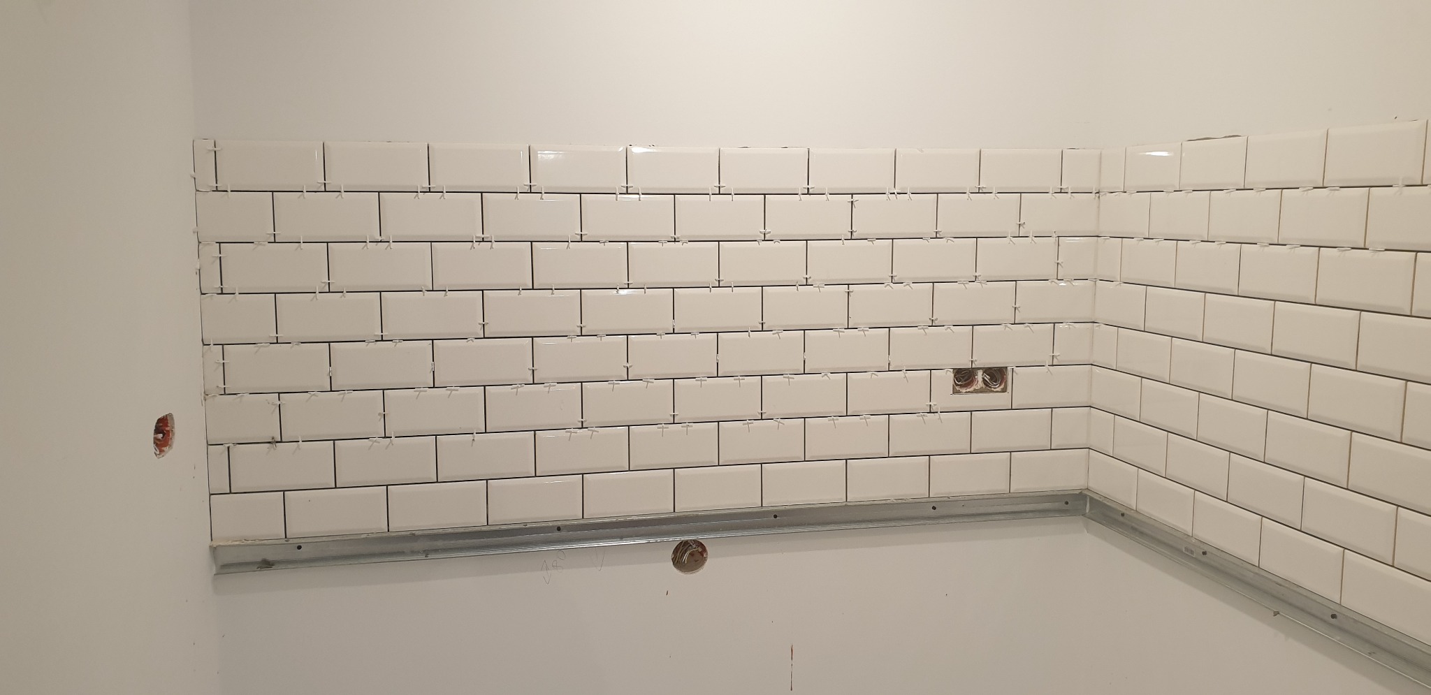Układanie białych, prostokątnych płytek na ścianie, z użyciem krzyżyków dystansowych i metalowej listwy podtrzymującej, widoczne puszki instalacyjne