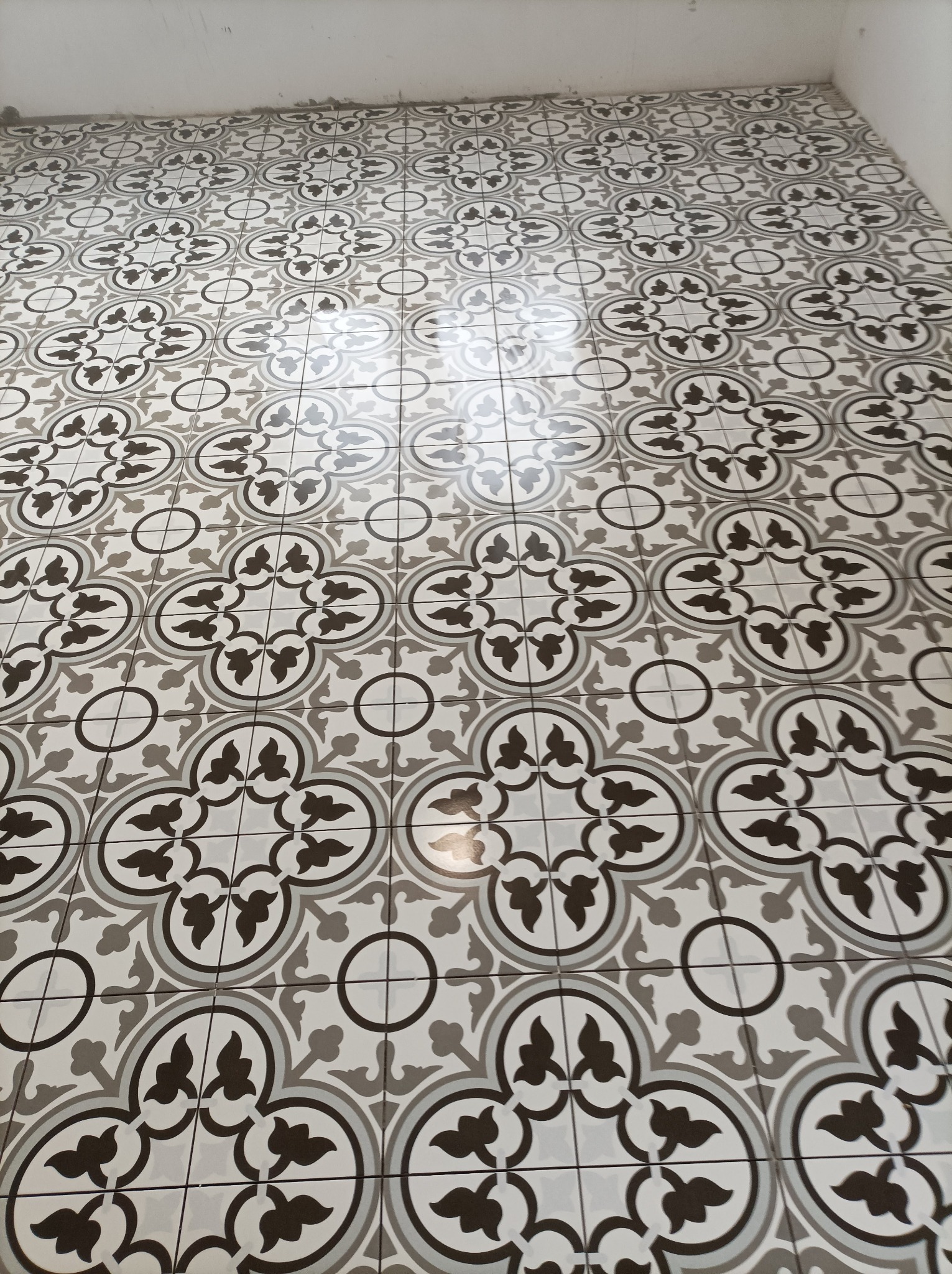 Ułożona podłoga z płytek ceramicznych w geometryczny wzór w odcieniach szarości i bieli, widoczne fugi i odbicie światła na powierzchni.