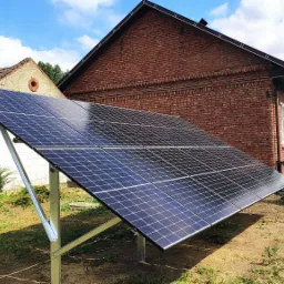 Panel słoneczny ustawiony na metalowej konstrukcji na trawniku przed ceglanym domem jednorodzinnym w słoneczny dzień.