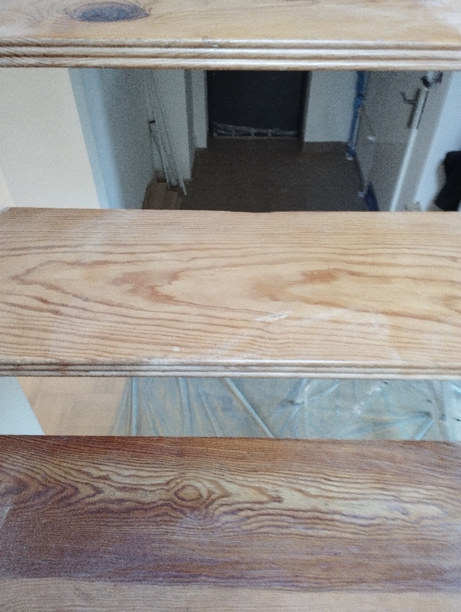 Drewniane schody w trakcie renowacji, widoczne trzy stopnie, z czego dolny jest już pomalowany bejcą, a dwa górne w stanie surowym, w tle zabezpieczone folią podłogi.