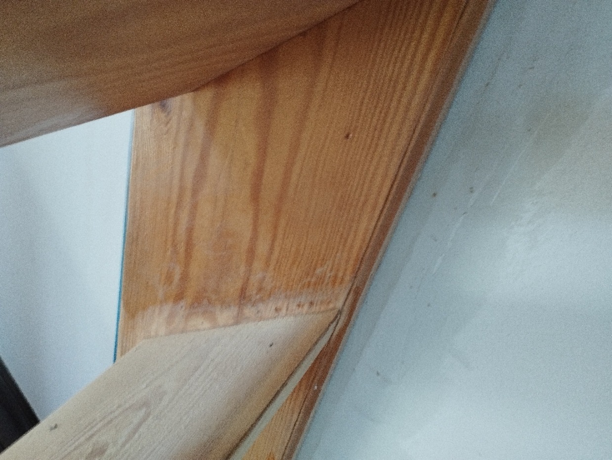 Fragment drewnianej konstrukcji w rogu łazienki, prawdopodobnie element obudowy wanny lub brodzika, przylegający do pomalowanej ściany.