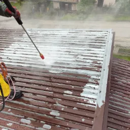 Czerwono-brązowy dach z blachy trapezowej, częściowo pokryty łuszczącą się białą farbą, czyszczony strumieniem wody pod ciśnieniem z czerwonej dyszy lance, widoczne narzędzia i fragment ulicy w tle.