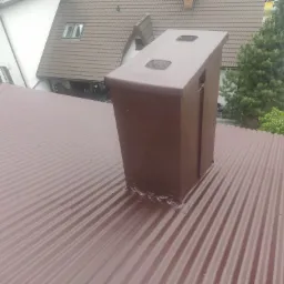 Ciemnobrązowy komin na metalowym dachu z falistej blachy, uszczelniony silikonem, z widocznymi elementami wentylacyjnymi na szczycie, w tle budynki mieszkalne i drzewa.