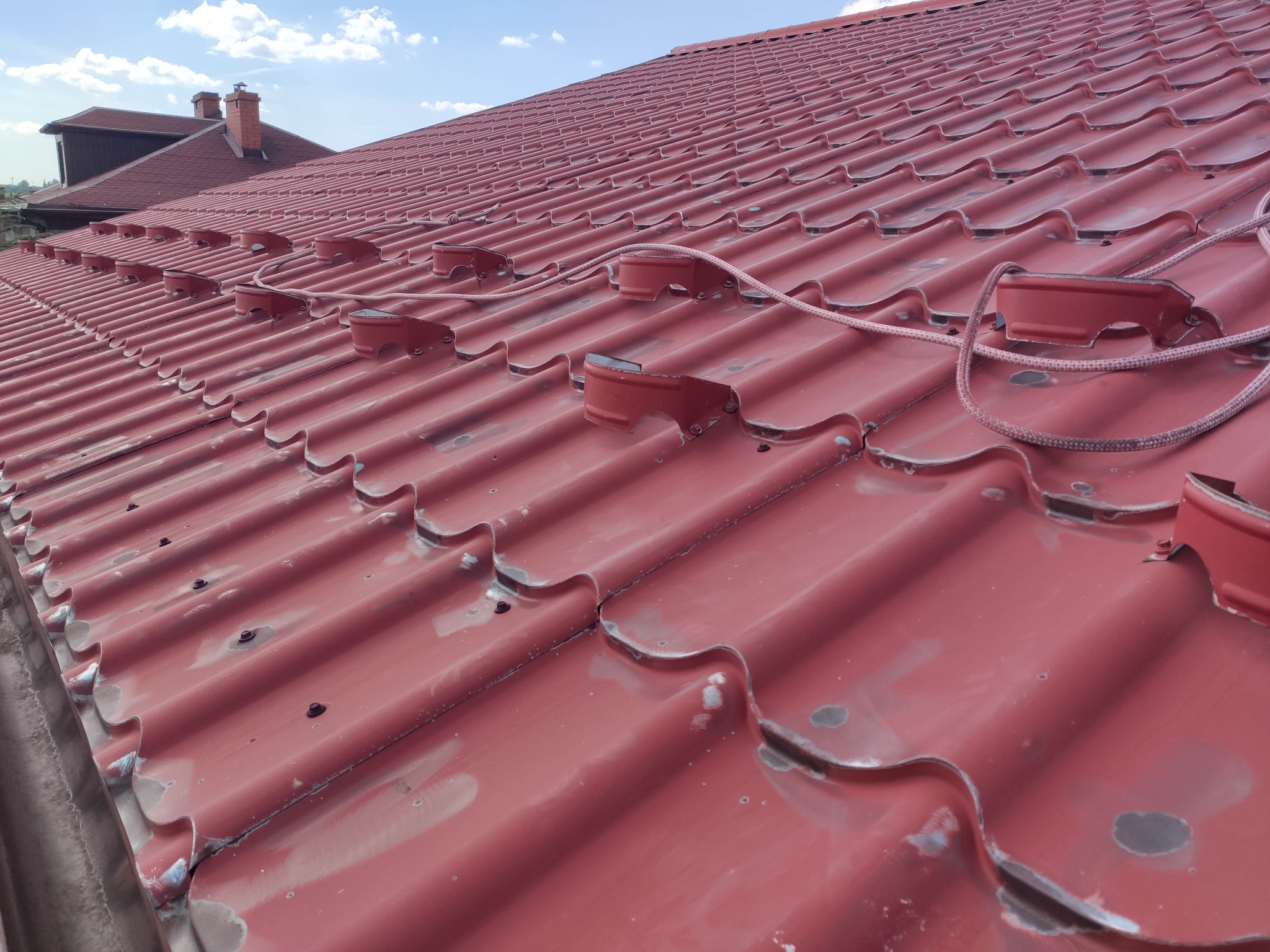 Czerwona blachodachówka z zamontowanymi uchwytami do zabezpieczenia linowego, widoczne punkty mocowania i ślady po wcześniejszych pracach.
