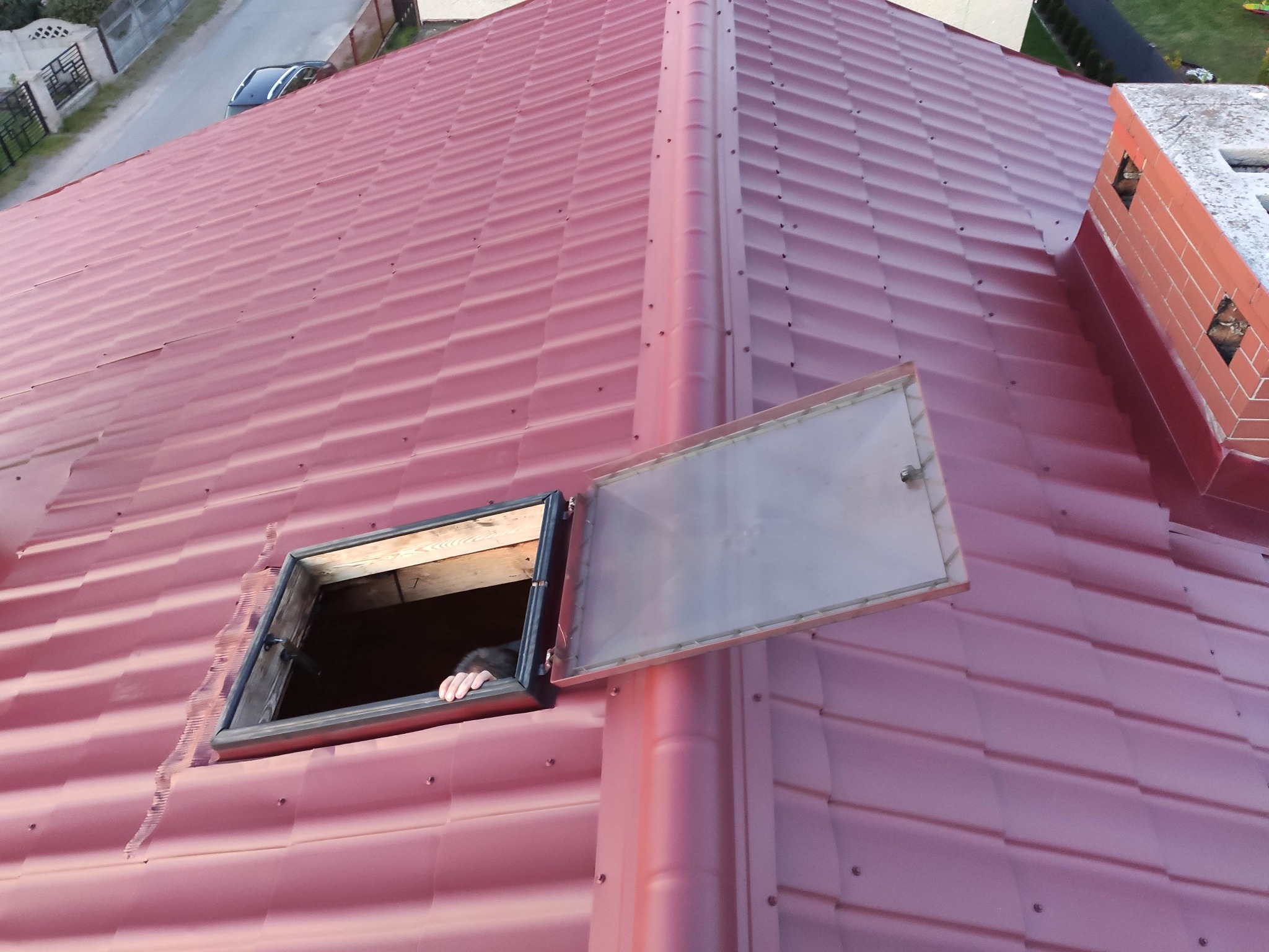 Czerwony dach z blachy trapezowej z otwartym oknem dachowym, z którego wystaje dłoń osoby. Widoczny komin na dachu i fragment ulicy w tle.
