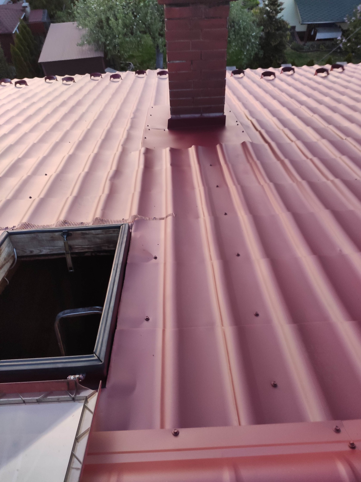 Czerwony dach z blachy trapezowej z kominem z cegły i otwartym oknem dachowym, widoczne mocowania blachy.