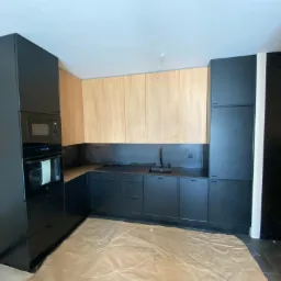 Częściowo ukończona kuchnia z czarnymi szafkami dolnymi i drewnianymi górnymi, zabezpieczona folią ochronną na podłodze, widoczne niedokończone podłączenia elektryczne.