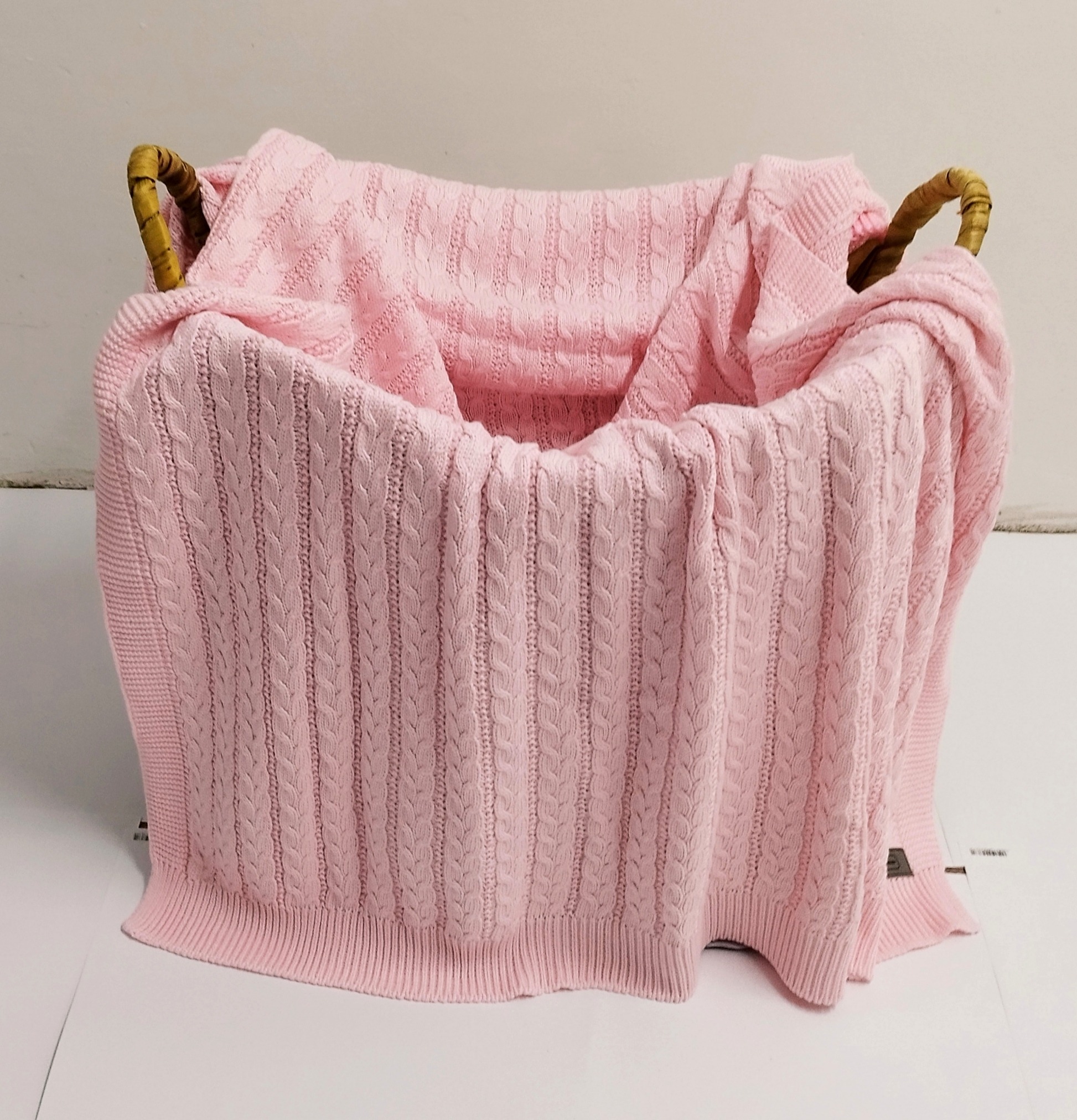 Różowy, dziergany kocyk dziecięcy z warkoczowym wzorem, ułożony w wiklinowym koszu z uchwytami, na białym tle.