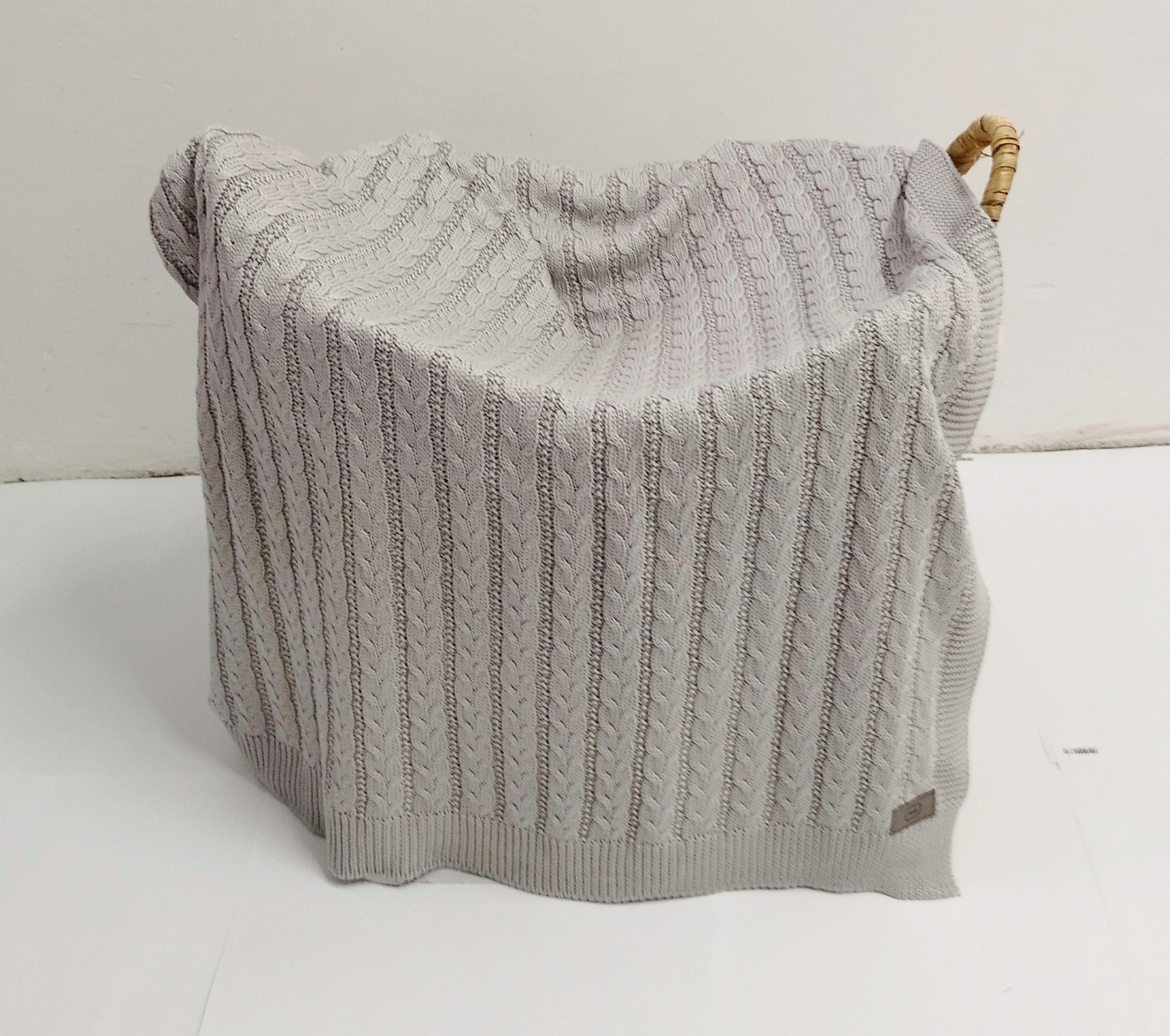 Szary, dziergany koc dziecięcy z wzorem w warkocze, częściowo ułożony w wiklinowym koszu na białym tle.