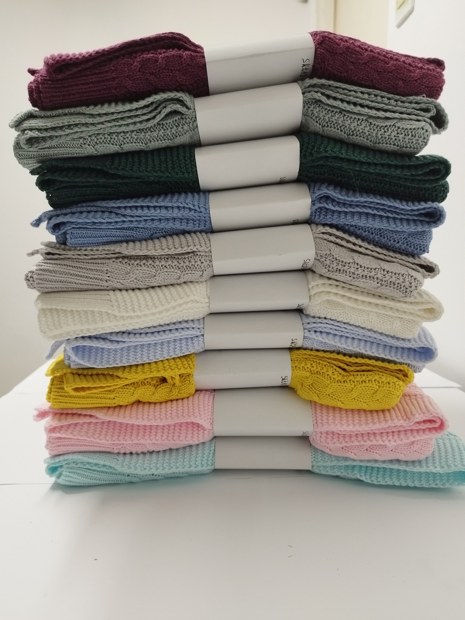 Stos dziesięciu zwiniętych, kolorowych, dzierganych kocyków dziecięcych z warkoczowym wzorem, przewiązanych białymi banderolami, ułożonych na białym stole.