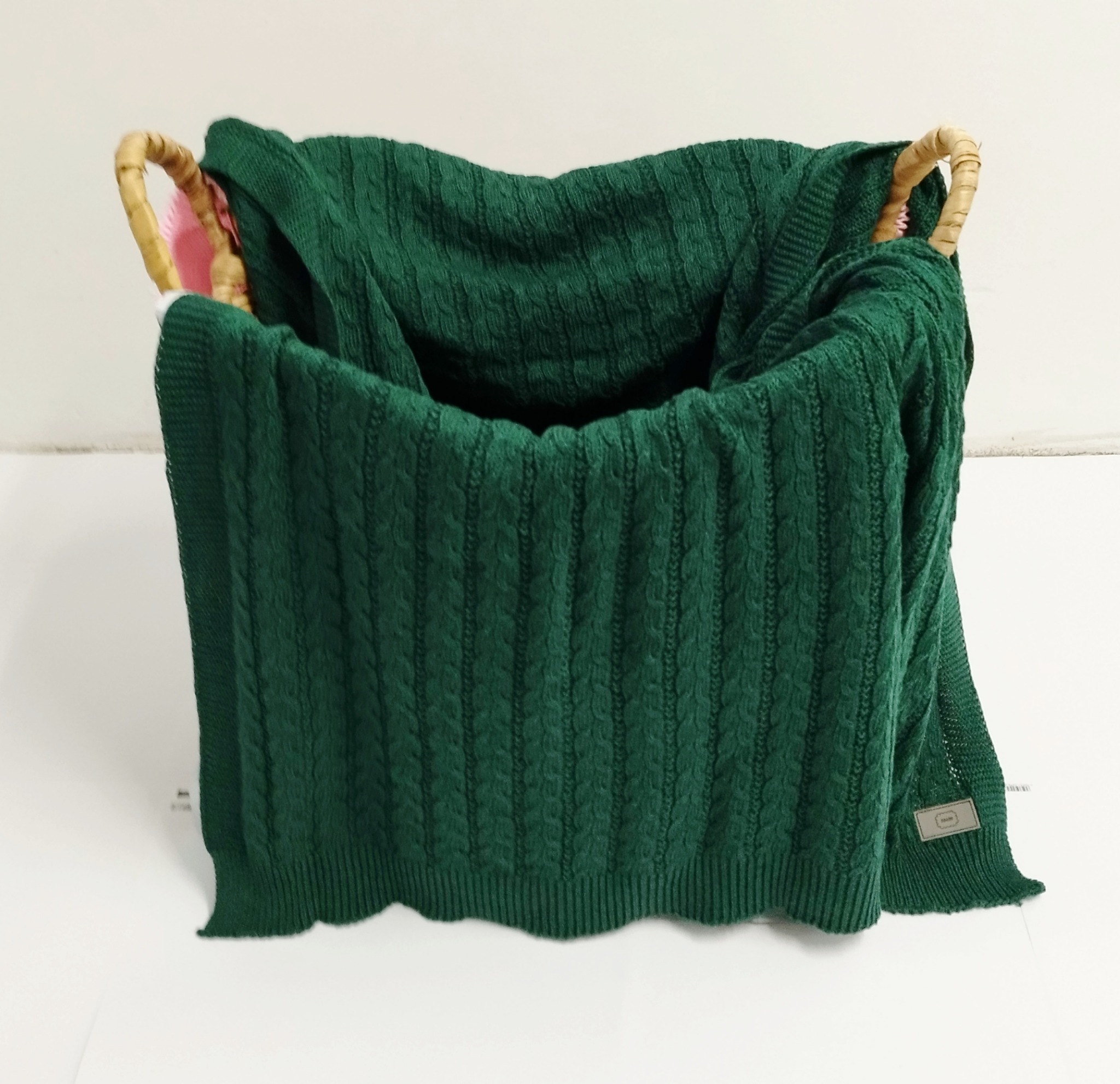 Ciemnozielony, dziergany koc z warkoczowym wzorem, umieszczony w wiklinowym koszu z bambusowymi uchwytami, na białym tle.