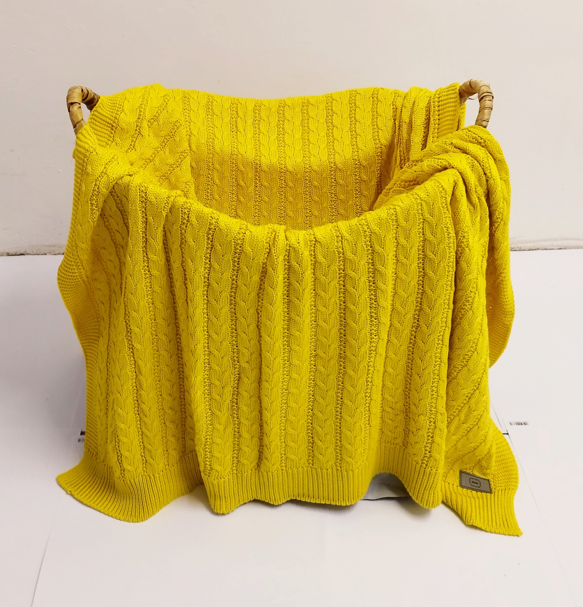 Żółty, dziergany koc z warkoczowym wzorem, ułożony w wiklinowym koszu na jasnym tle.