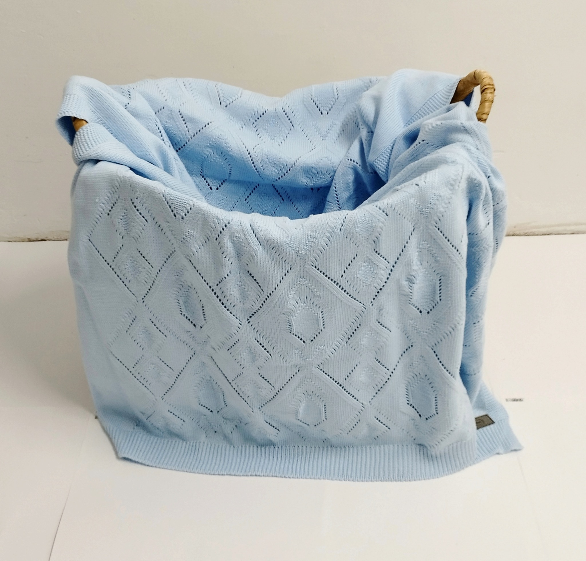Jasnoniebieski, ażurowy kocyk dziecięcy z geometrycznym wzorem, ułożony w wiklinowym koszu.