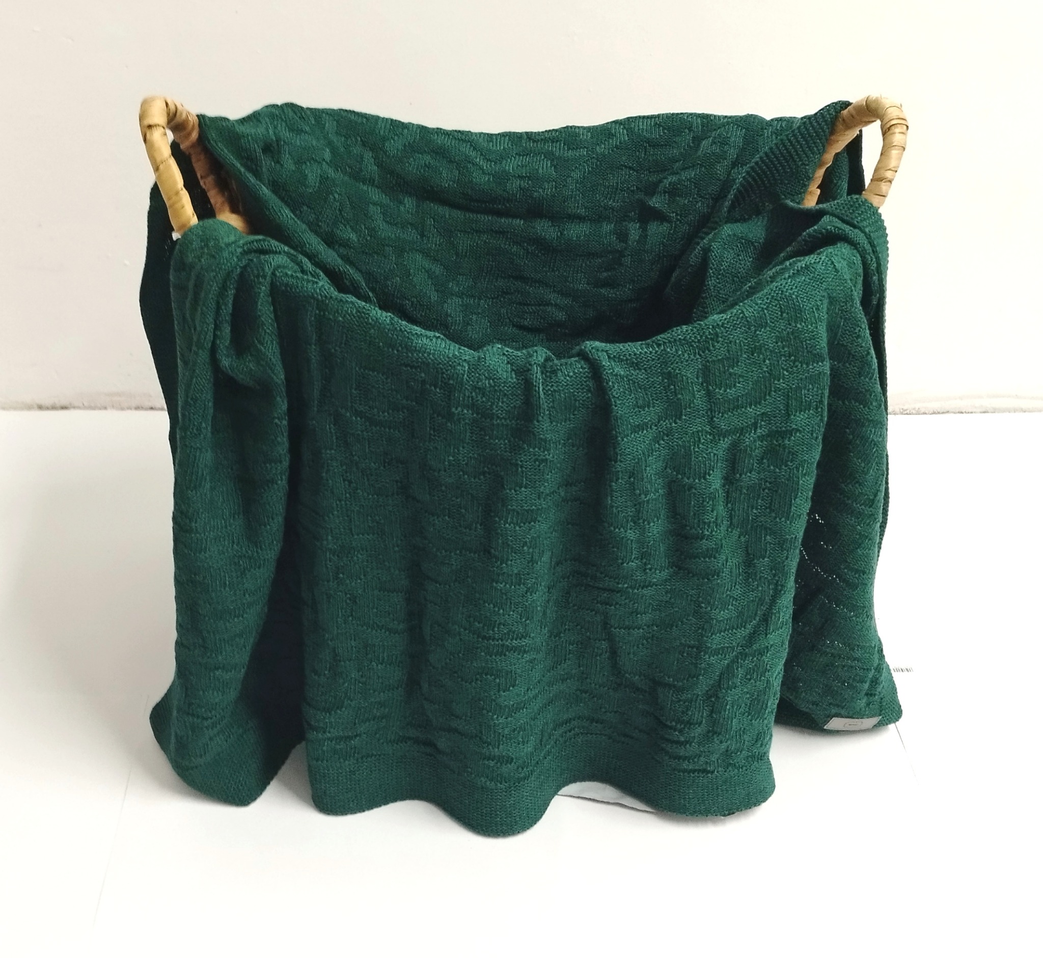 Szmaragdowy, dziergany koc w wiklinowym koszu z uchwytami, prezentowany przez firmę z Elbląga.