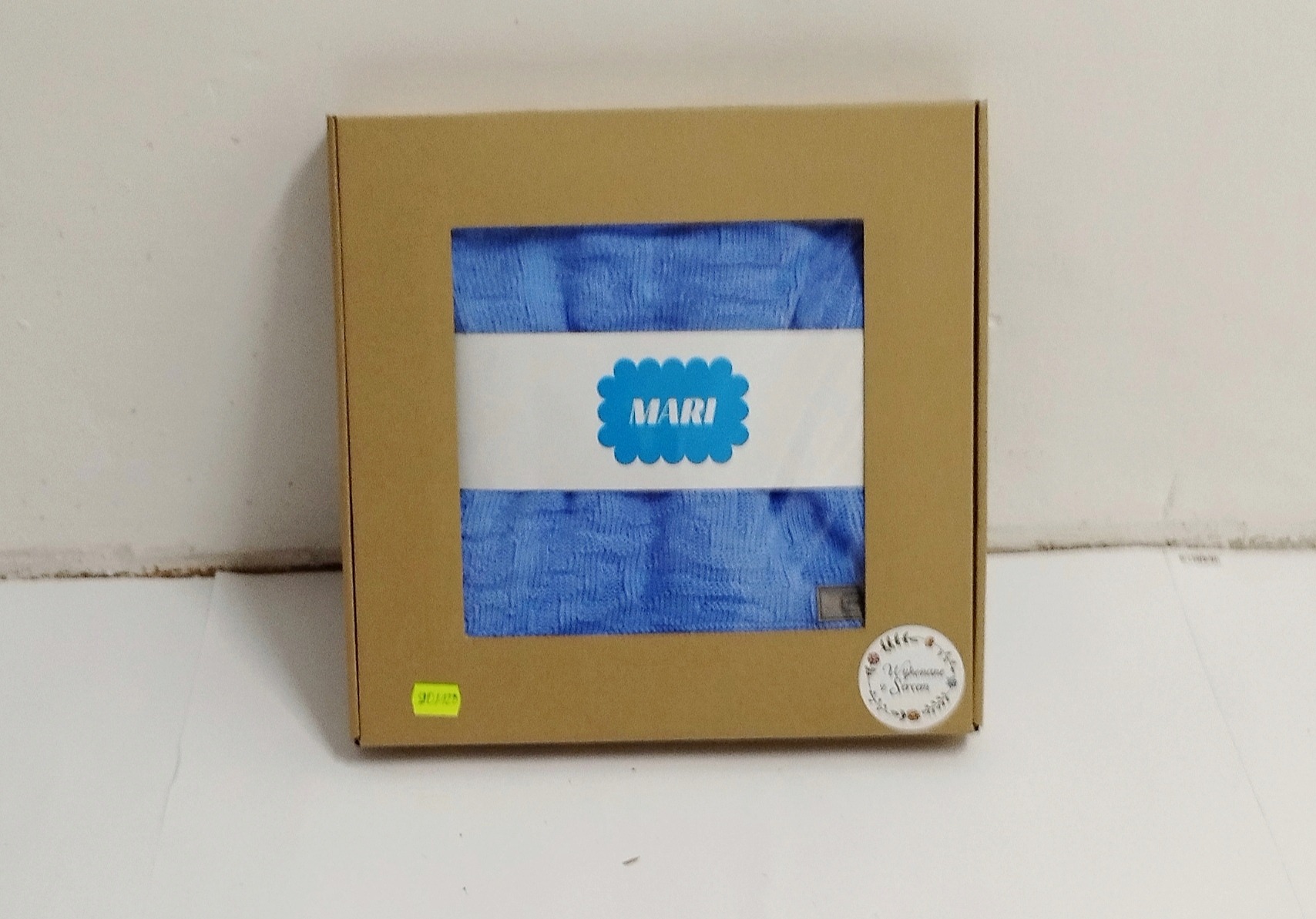 Jasnoniebieska, dziergana chusta dziecięca z imieniem 'MARI' na etykiecie, zapakowana w kartonowe pudełko z naklejką z logo firmy.