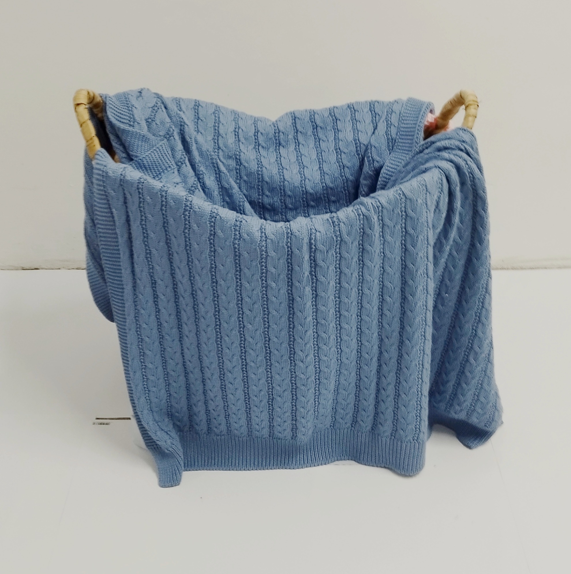 Jasnoniebieski, dziergany kocyk z warkoczowym wzorem, ułożony w wiklinowym koszu na białym tle.