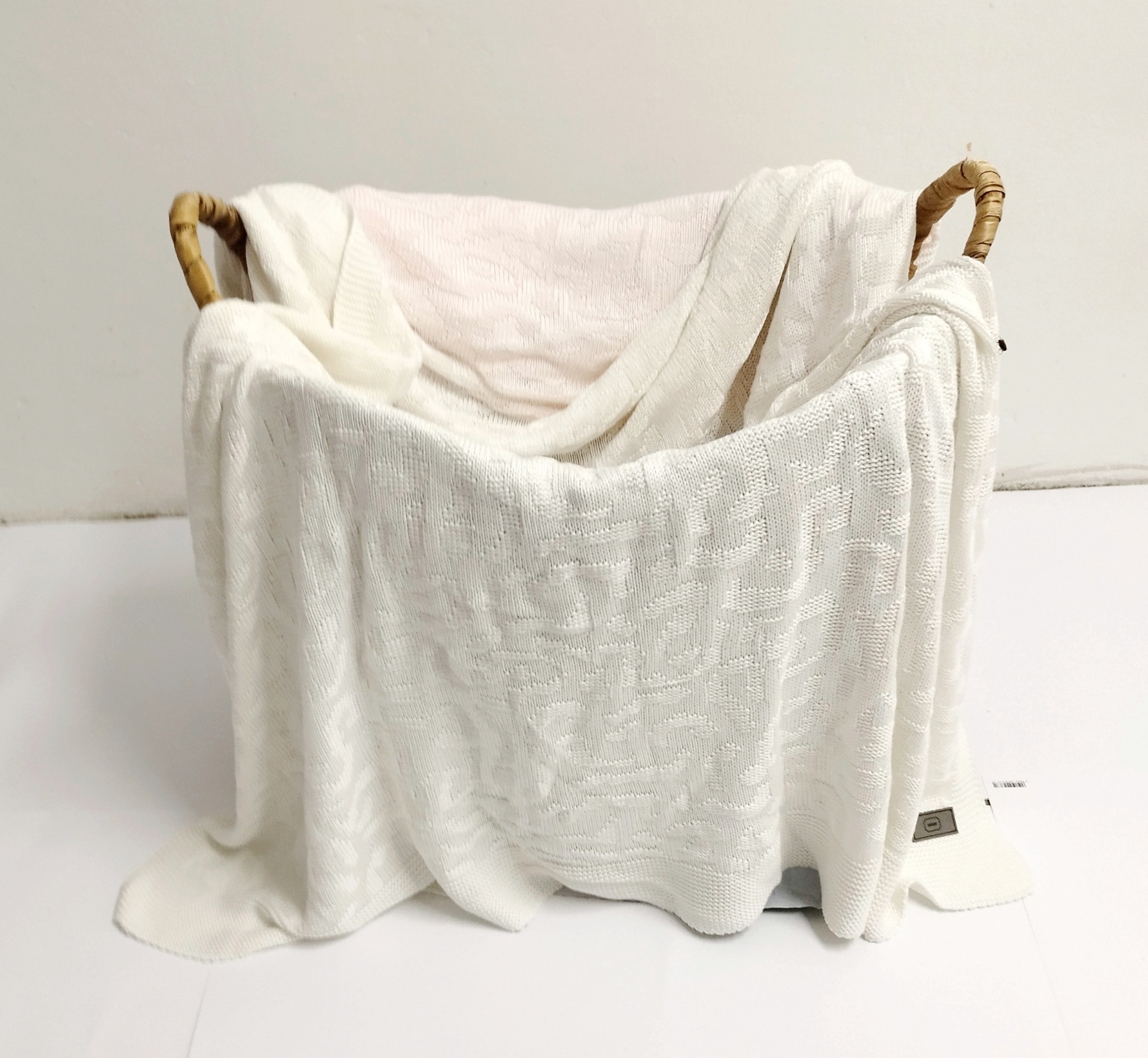 Kosz wiklinowy z kremowym, dzierganym kocykiem dziecięcym z subtelnym wzorem, widoczna metka.