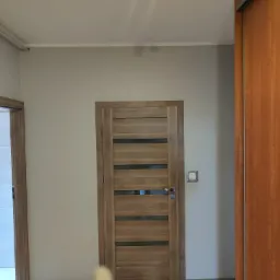 Wnętrze pokoju z drewnianą, przeszkloną poziomymi paskami, drzwiami wewnętrznymi, szarą ścianą, widocznym włącznikiem światła i fragmentem brązowej szafy.