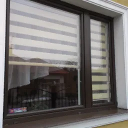 Brązowe okno z roletami dzień noc, widoczne z zewnątrz, z refleksami otoczenia i pudełkiem klocków Lego Friends na parapecie.