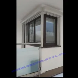 Zabudowany balkon z oknami z brązowymi ramami i zewnętrznymi roletami w kolorze białym, widok z zewnątrz, częściowo widoczna szklana balustrada.