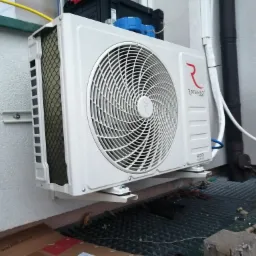 Biała jednostka zewnętrzna klimatyzatora marki Rotenso zamontowana na ścianie budynku, widoczna kratka wentylacyjna i elementy instalacji.