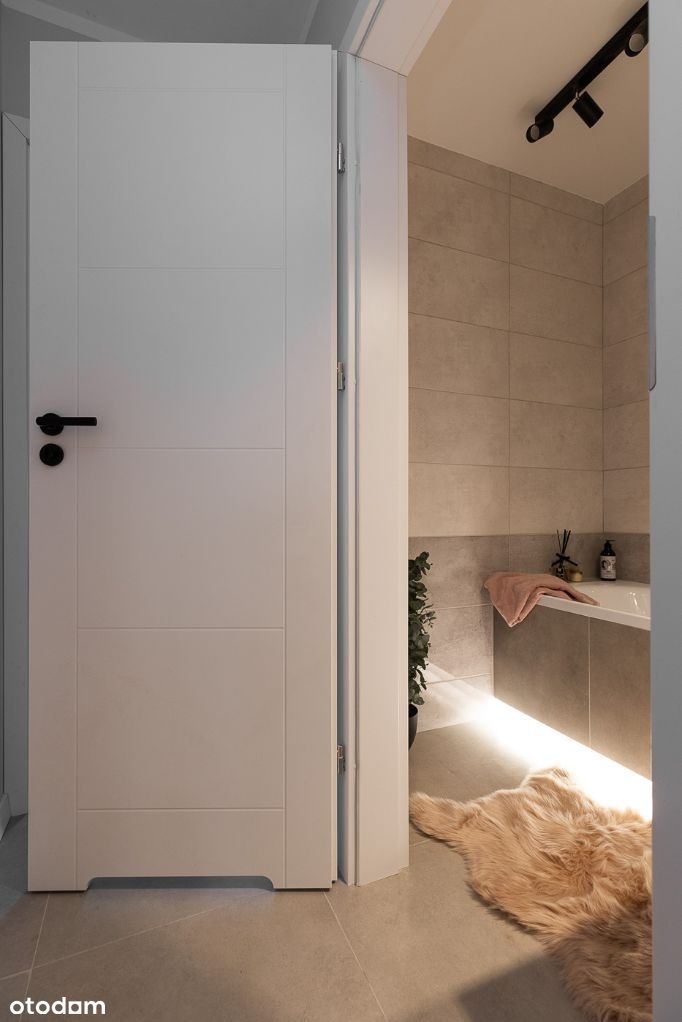 Otwarta biała, panelowa drzwi do łazienki z beżowymi płytkami, podświetloną wanną, dywanikiem imitującym skórę owczą i czarnym oświetleniem punktowym na suficie.