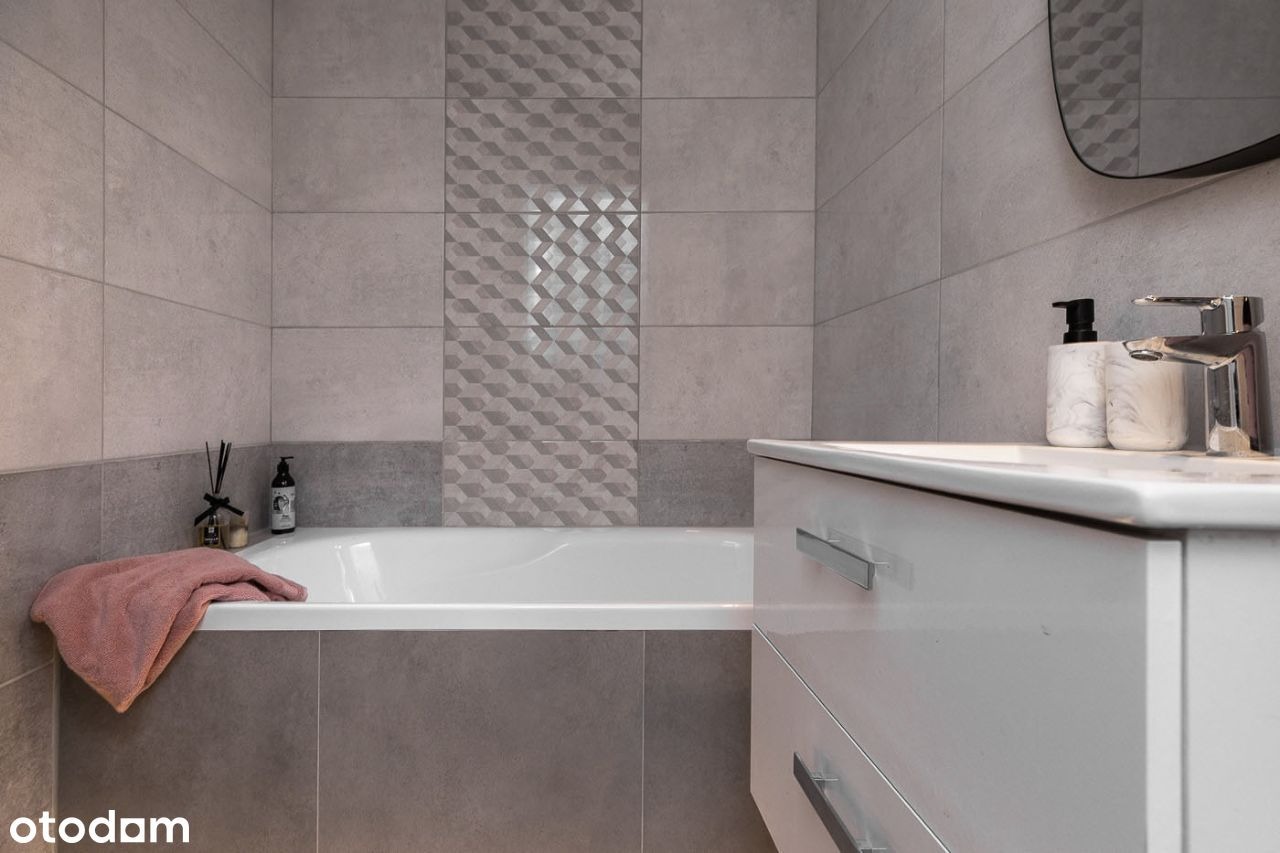 Nowoczesna łazienka w odcieniach szarości z geometrycznym wzorem na płytkach, białą wanną, umywalką z szafką i akcesoriami łazienkowymi.