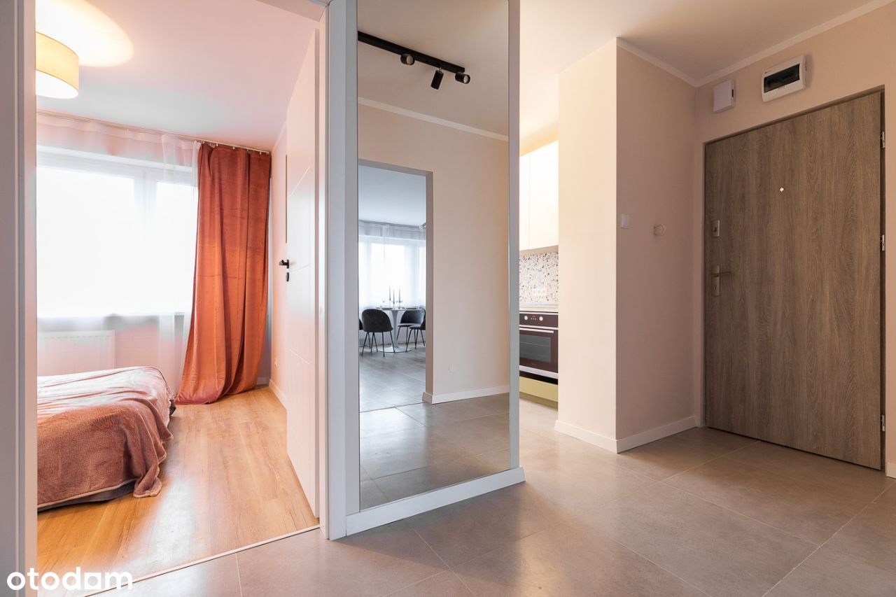 Widok na wnętrze mieszkania z korytarza: sypialnia z zasłoniętym oknem, lustro w białej ramie odbijające jadalnię i fragment kuchni, oraz drzwi wejściowe z szarą okleiną.