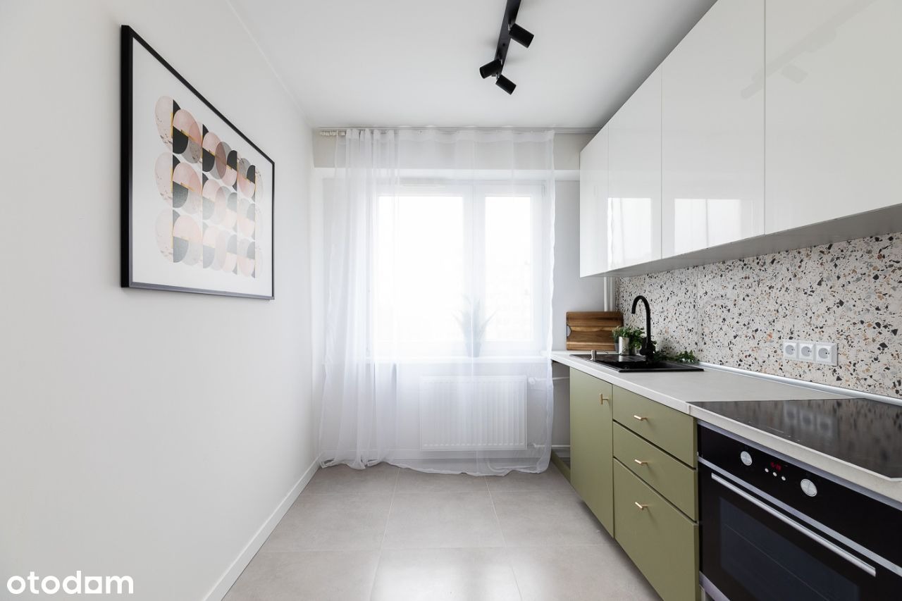 Nowoczesna kuchnia z zielonymi szafkami, białymi górnymi szafkami, mozaikową ścianą nad blatem i geometrycznym obrazem na ścianie.