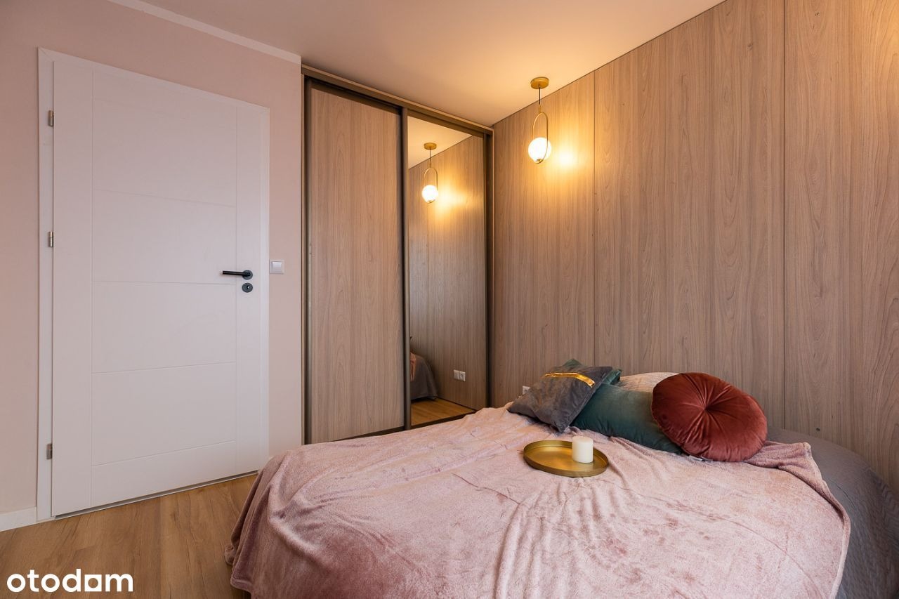 Nowoczesna sypialnia z drewnianą ścianą, przesuwną szafą z lustrem i białymi drzwiami, oświetlona wiszącymi lampami i z łóżkiem nakrytym pluszową narzutą w odcieniach różu i szarości.