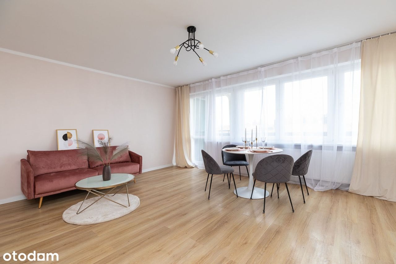 Jasne wnętrze mieszkania z różową ścianą, welurową sofą, okrągłym stolikiem kawowym, okrągłym stołem jadalnym z szarymi krzesłami i dużym oknem z prześwitującymi zasłonami.