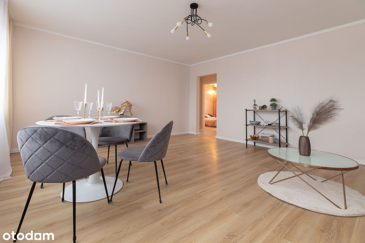 Jasne wnętrze pokoju dziennego z okrągłym stołem, szarymi krzesłami, drewnianą podłogą i dekoracjami, widok na sypialnię w tle.