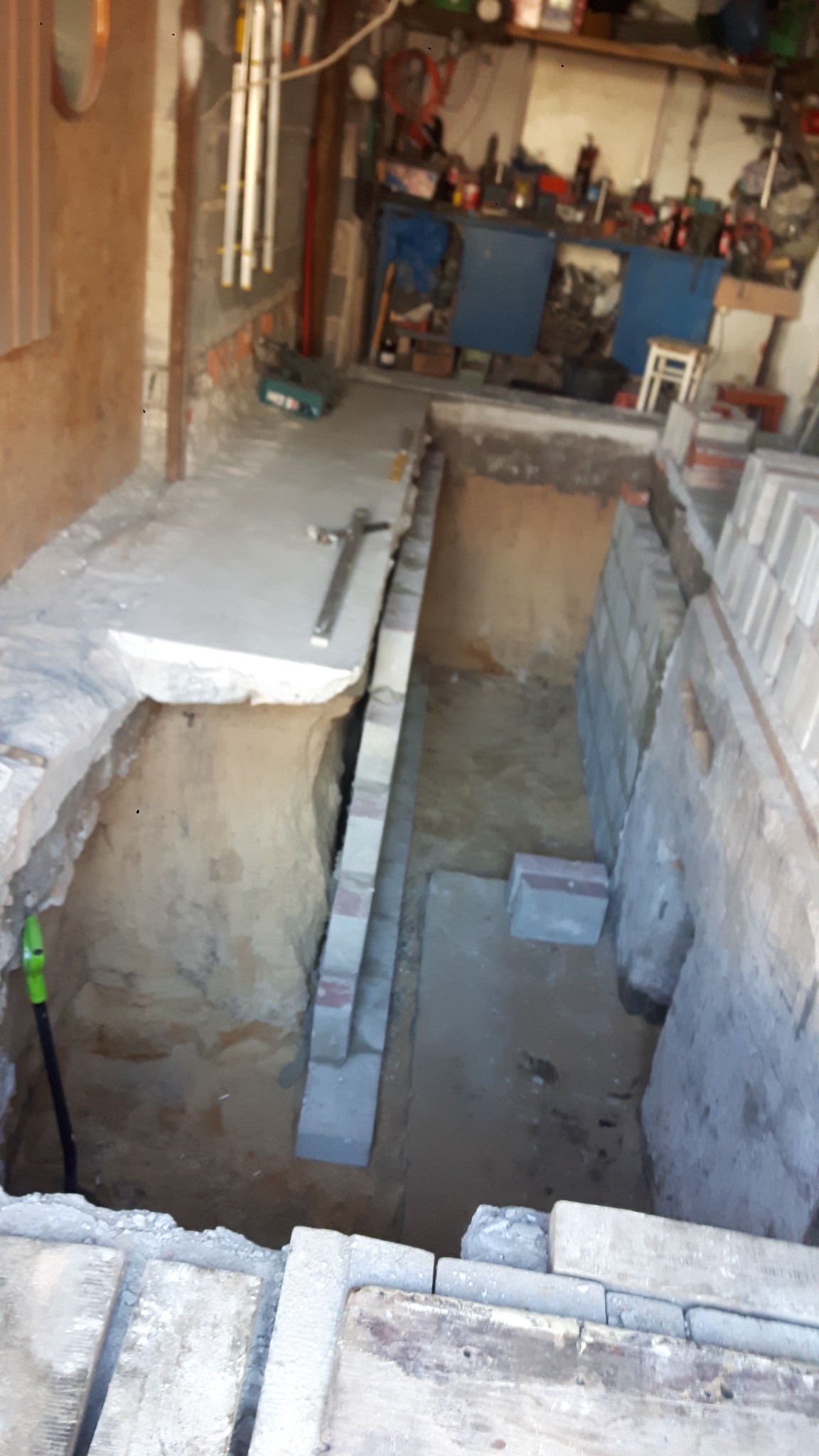 Wykop wewnątrz budynku z częściowo ułożonymi bloczkami fundamentowymi, widoczna łopata z zielonym uchwytem i narzędzia budowlane w tle.