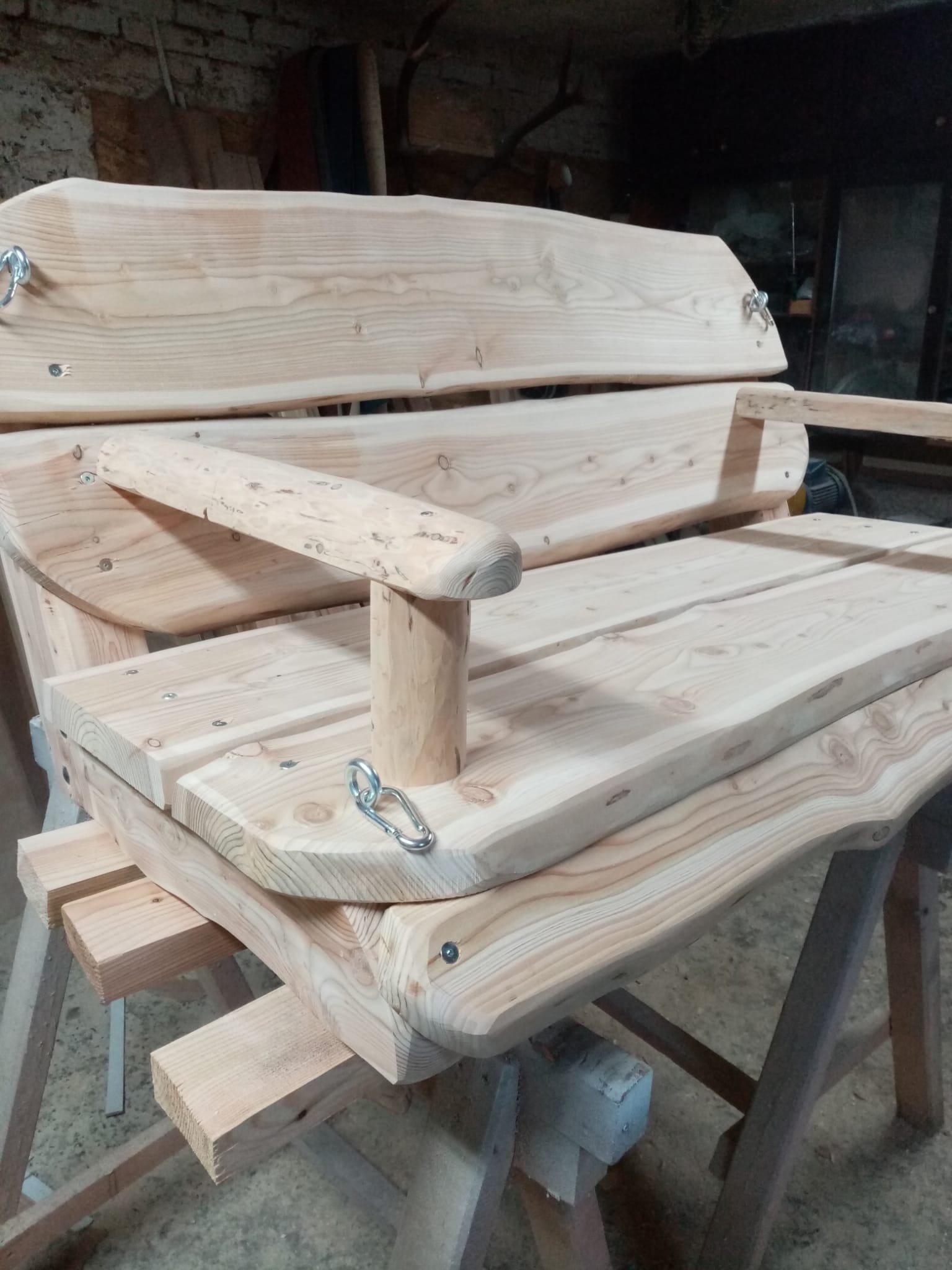 Surowy, drewniany bujak ogrodowy na drewnianych kozłach, z widocznymi śrubami i karabińczykami, w warsztatowym otoczeniu.