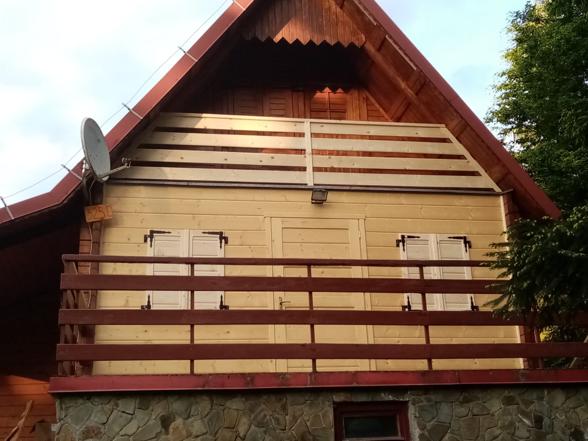 Fasada drewnianego domu z balkonem, widoczne jasne deski, ciemnobrązowa balustrada, kamienna podstawa i antena satelitarna.