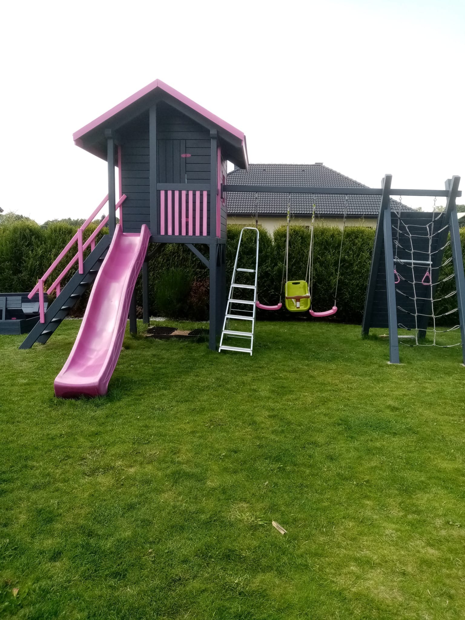 Drewniany plac zabaw dla dzieci w odcieniach szarości i różu, z domkiem na podeście, zjeżdżalnią, schodami, drabinką, huśtawką i ścianką wspinaczkową na tle zielonego trawnika i żywopłotu.