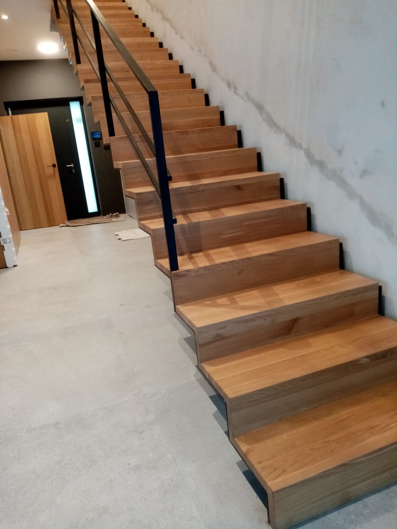 Nowoczesne schody drewniane z czarną balustradą, widok z dołu, szare płytki podłogowe i drzwi w tle.