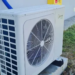 Biała zewnętrzna jednostka pompy ciepła Midea zamontowana na betonowym fundamencie z czarnymi podkładkami antywibracyjnymi, widoczna żółta skrzynka gazowa na ścianie budynku.