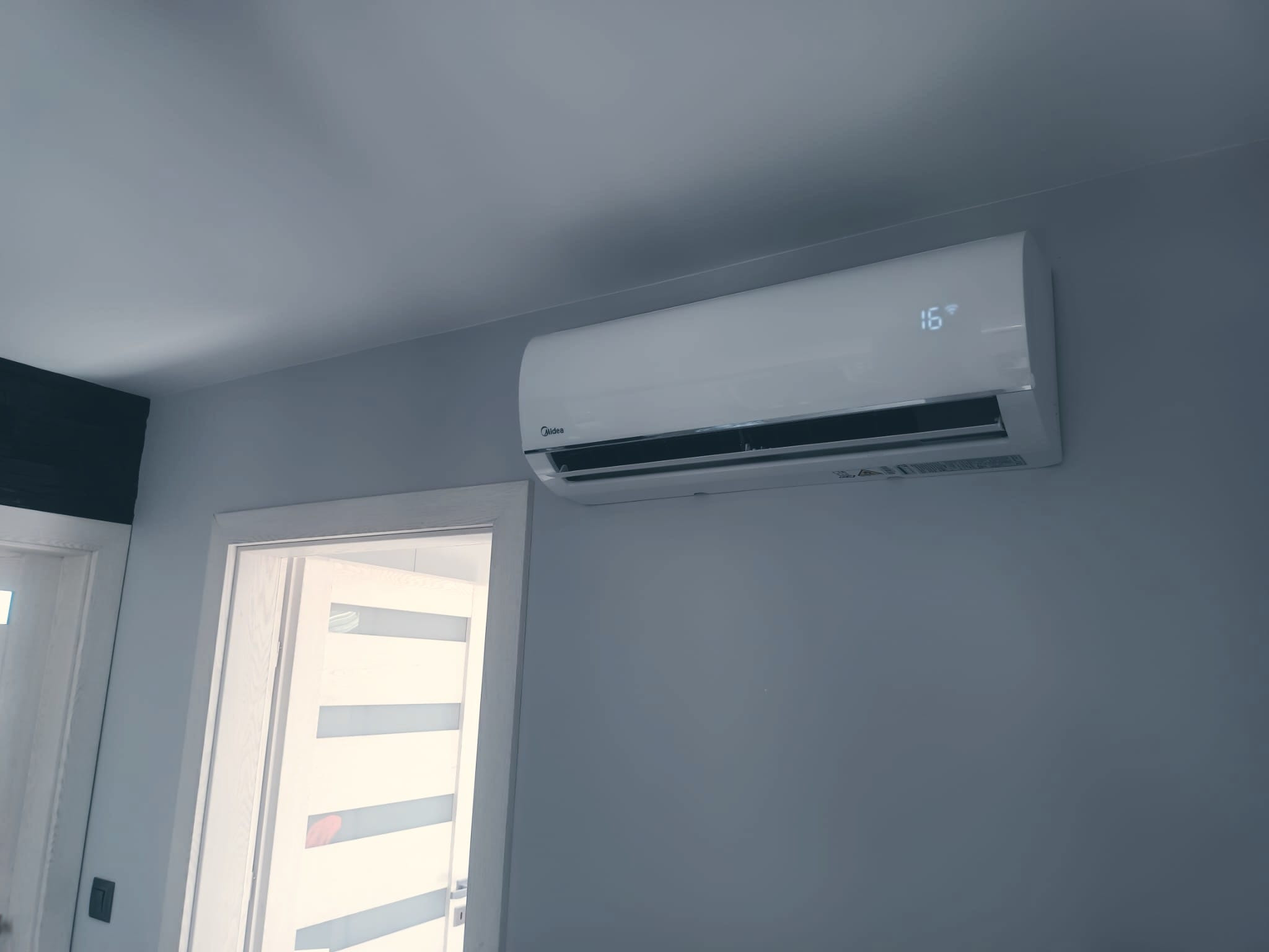 Biały klimatyzator Midea zamontowany na szarej ścianie, wyświetlający temperaturę 16 stopni, widoczny fragment białej ościeżnicy drzwi z lewej strony.