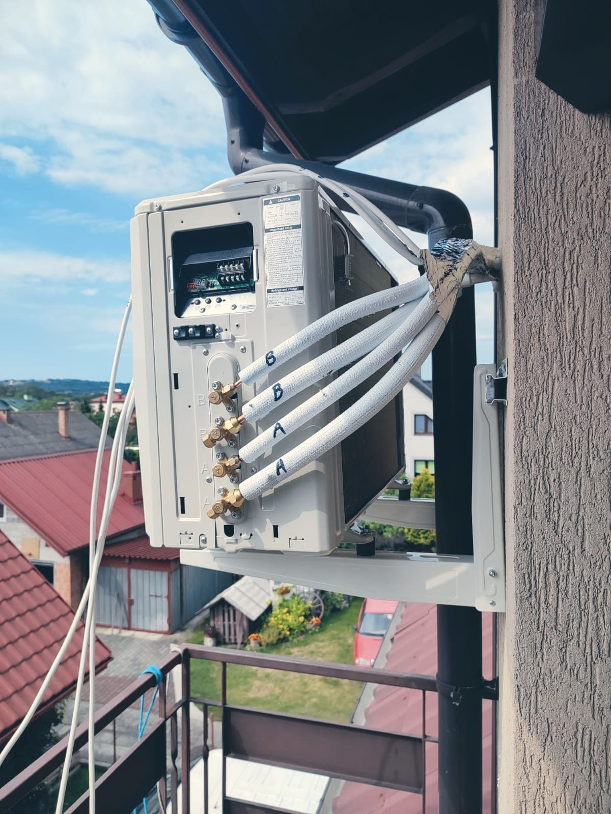 Zewnętrzna jednostka klimatyzacji zamontowana na balkonie, widoczne podłączenia rur chłodniczych z oznaczeniami A i B, na tle zabudowań mieszkalnych i błękitnego nieba.