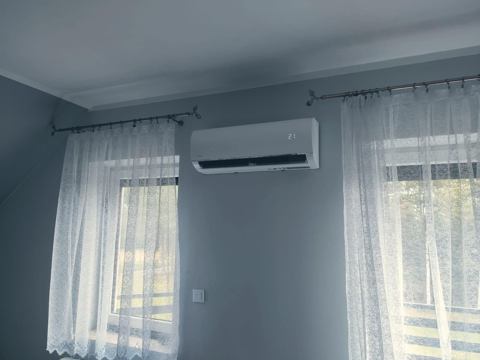 Biała klimatyzacja ścienna Midea z wyświetlaczem temperatury 21 stopni Celsjusza, zamontowana na szarej ścianie pomiędzy oknami z białymi, koronkowymi zasłonami i karniszami z motywem liści.