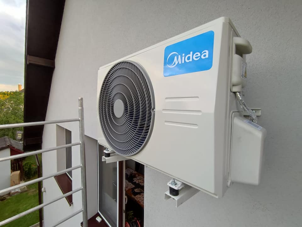 Biała jednostka zewnętrzna klimatyzacji Midea zamontowana na wspornikach na ścianie budynku, widok z balkonu z metalową barierką.